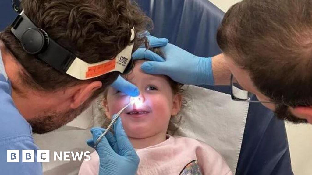 Peyton Handley - smiling and lying on a hospital bed as two doctors examine her nose, shining a torch and inserting tweezers in her nostril
