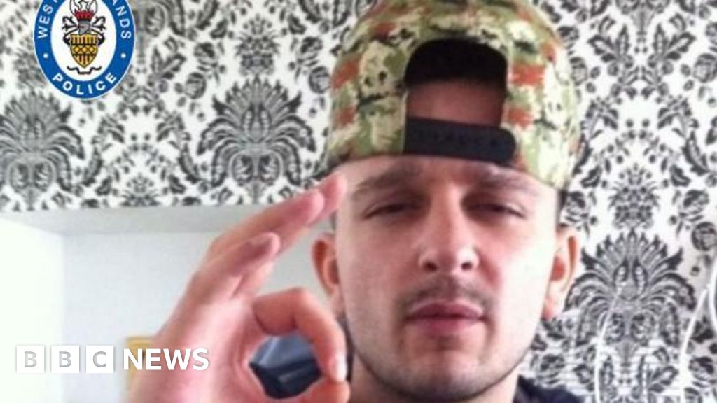 A man  wearing a khaki baseball cap worn backwards looks into the camera. he has a bit of a moustache and is holding his thumb and forefinger together. He is inside a property which has a black and white wallpaper pattern behind him.