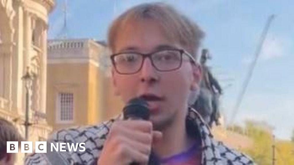 A man with blonde hair and glasses who is wearing a white and black scarf over his shoulders speaks into a microphone