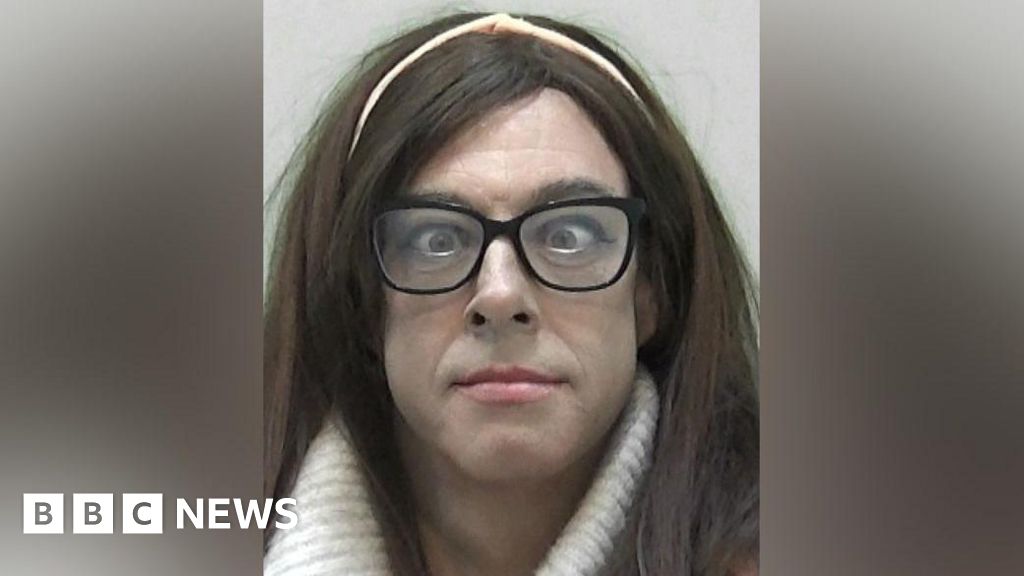 Mugshot of Natalie Wolf, formerly known as Ryan Haley, who has thick long brown hair with a pink hairband in, with slight make up and pink lipstick and black-framed glasses over a pair of cross-eyed eyes.