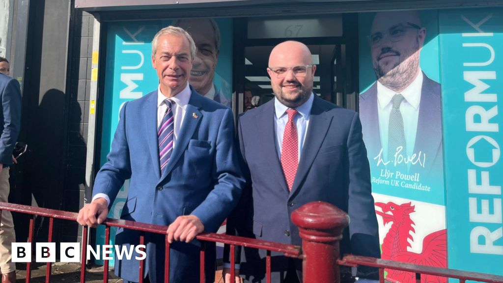 Nigel Farage and Llŷr Powell standing behind a red railing in front of Reform's office in Caerphilly. Both men are wearing blue suits.