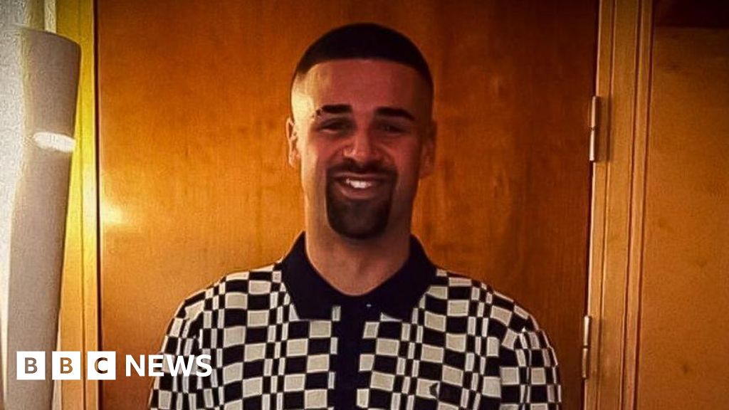 Tariq Evans, who has a shaved head and eyebrow, has a dark beard and moustache and is wearing a black and white check polo shirt