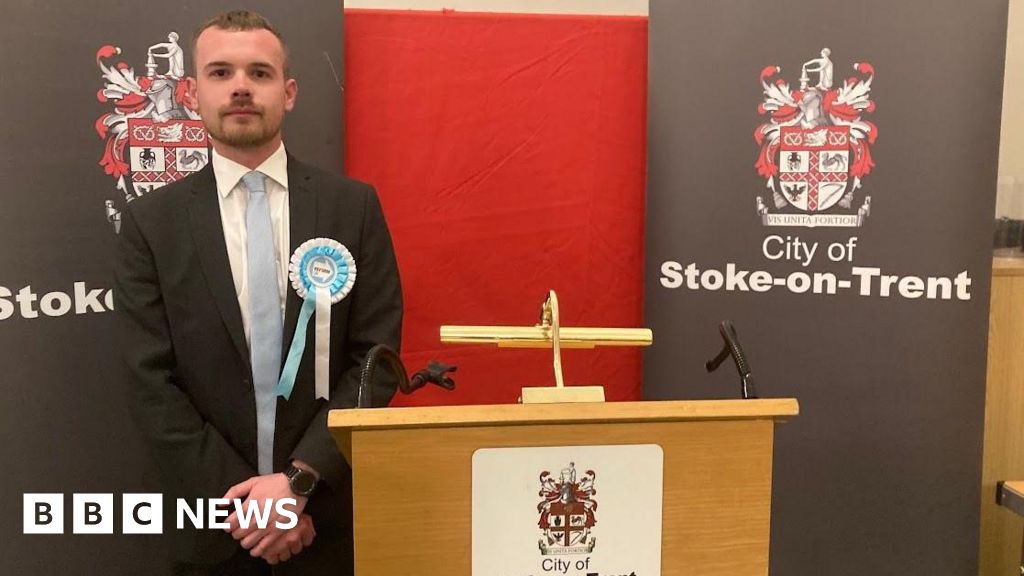 A man stands next to a podium at a by-election count. He has his hands clasped in front of him and he's wearing a blue and white Reform rosette. 