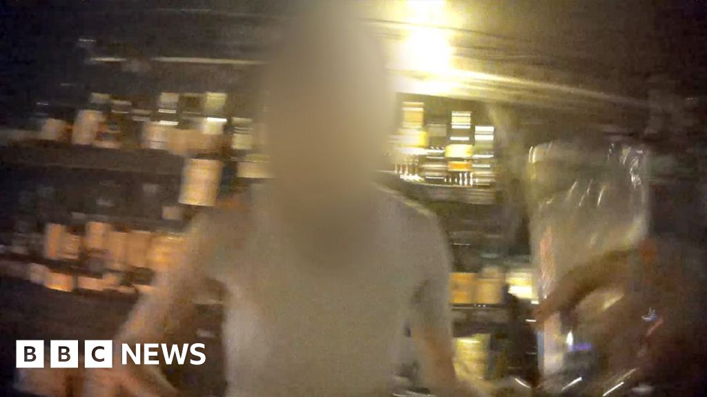 Blurred image of a man at a bar at night taken with a secret camera. In the foreground a hand is holding a glass, in the background there are bottles on shelves.