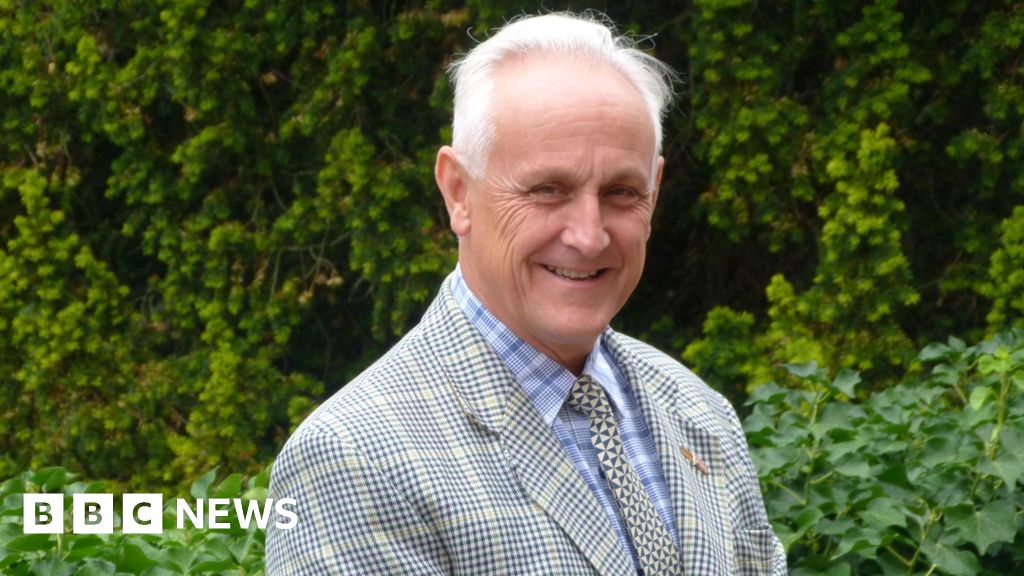 A man with white hair is wearing a checked suit, a shirt and tie and is stood smiling in front of greenery