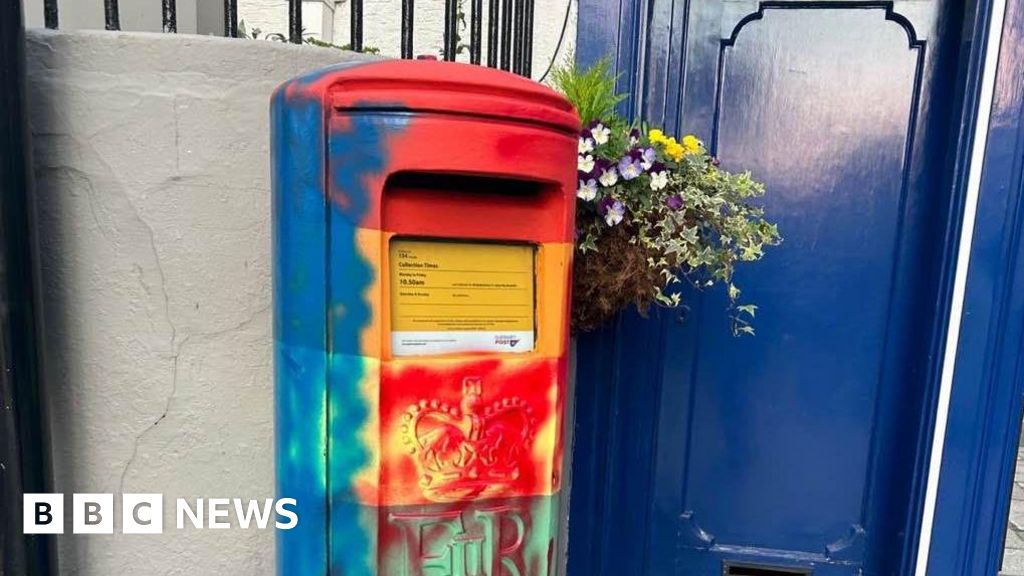 The photograph shows a Guernsey postbox that had been painted in the colours of the rainbow. There is now blue spray paint up the left side of it and red spray paint up the right side of it and on the front.  