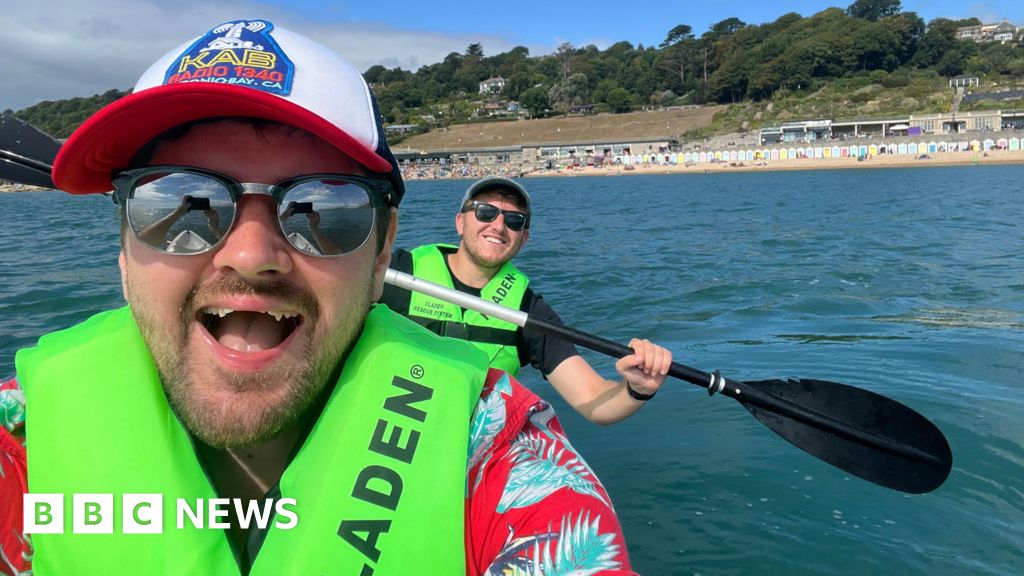 Two men in a kayak. The man closest to the camera is taking a selfie with another man holding a paddle horizontally in the background. Both are smiling and wearing sunglasses and green life vests. In the background the sea, shore and trees on the hills...
