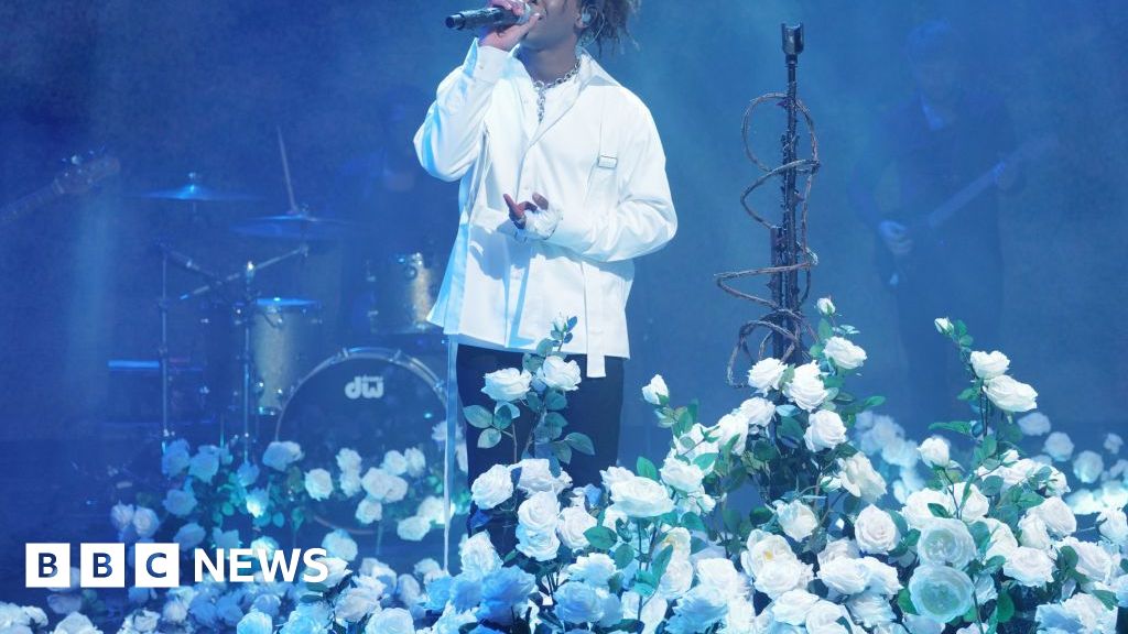 Wearing a white jacket and surrounded by white roses, D4vid sings into a microphone