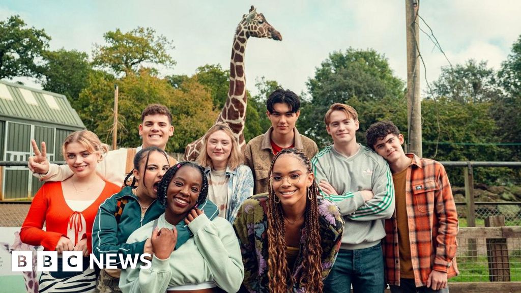 The cast of Heartstopper pose happily for a photo with a giraffe looming behind them