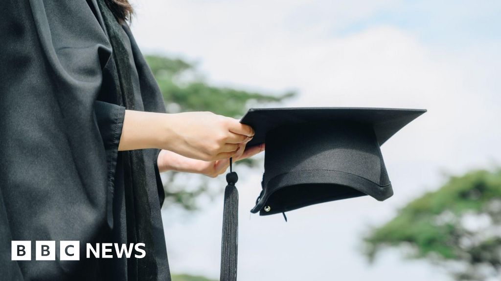 A person in a graduation gown holding a graduation cap with a tassel, standing outdoors with trees and a cloudy sky in the background.