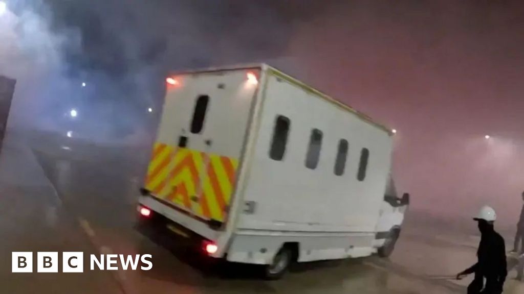 A screenshot of the police van seen on the Elbit Systems UK site. The van is a large white vehicle with small windows and a chevron pattern across the back. A figure wearing black with a white helmet can be seen in the lower right corner of the image h...