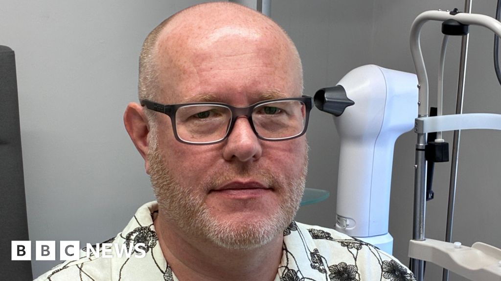 A man wearing glasses and a white shirt with black flowers on it. He is in a room at an optician surgery.