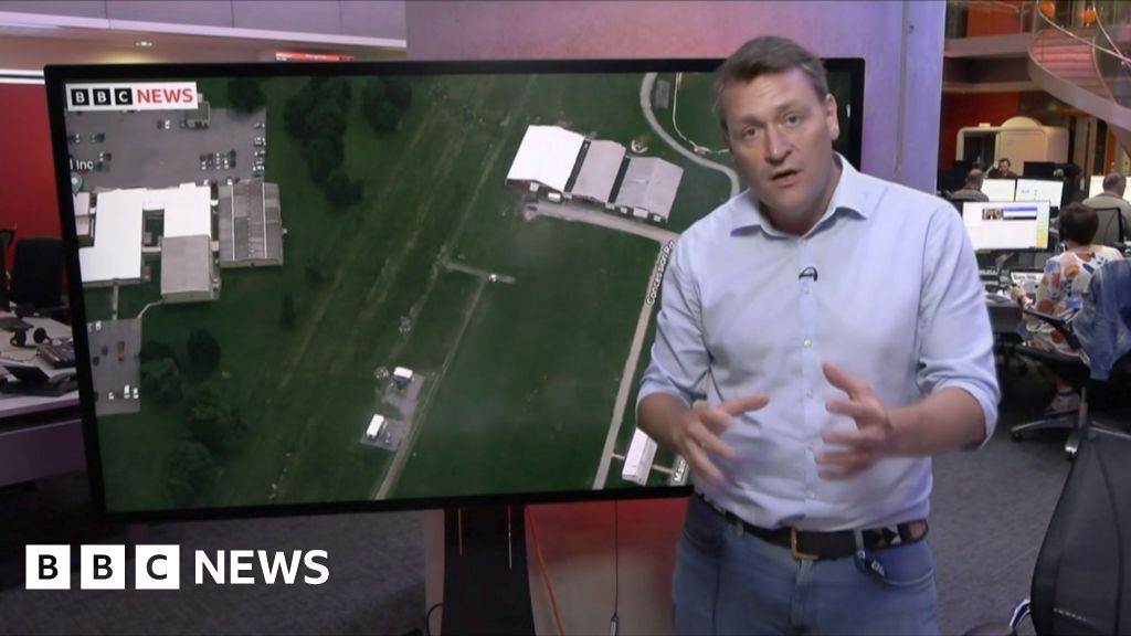 Joe Inwood stands in front of a TV screen showing an aerial of a green field with several buildings in it, the site of the attempted assassination 