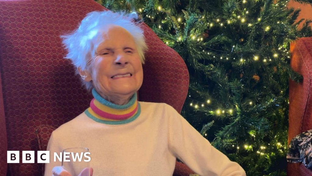Molly, who is wearing a rainbow coloured jumper, is sitting in a red armchair and smiling. She is holding a champagne glass.