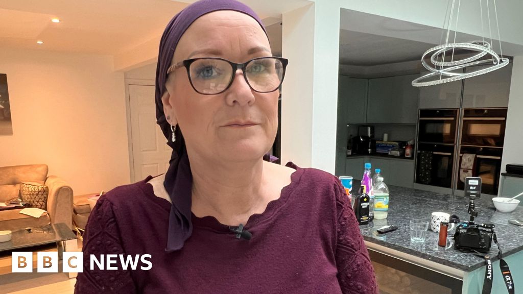 Paula Hudgell wears black framed glasses, a maroon top and matching headscarf as she sits in the kitchen of her home, looking directly into the camera.