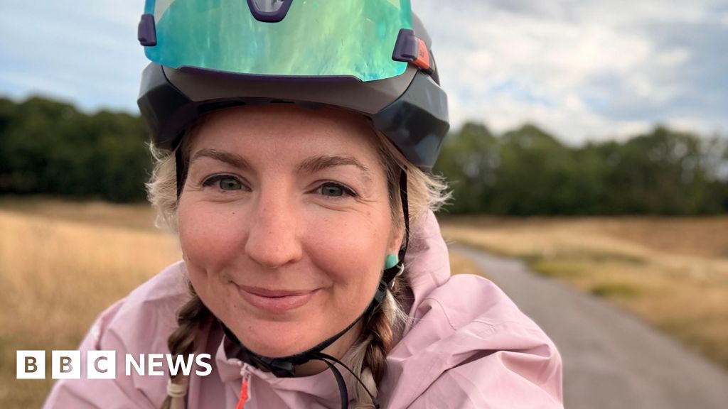 Claire Sharpe wears a cycling helmet and pink jacket has taken a selfie whilst riding her bike through a rural area. She has blonde hair and is smiling.