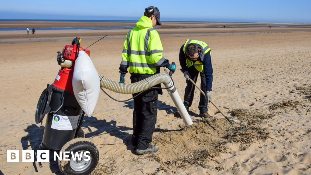 Two men, operating a vacuum on the beach. They are both wearing high visibility jackets. One is holding the vacuum, whilst the other uses a tool to dislodged sand.