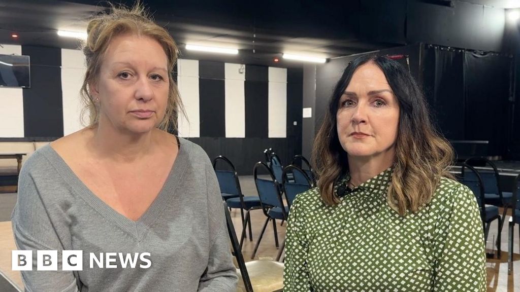 A woman wearing all-grey with blonde hair tied back sitting next to another woman wearing a green patterned blouse. They are inside.