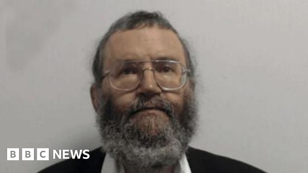 Dr Wayne Davis with brown eyes, grey short hair and long grey beard and glasses in photograph taken by police in custody