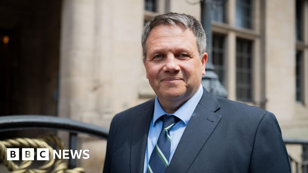 Andrew Mynors, a man wearing a dark suit with a blue shirt and striped tie. He is standing outside the County Buildings in Stafford, a