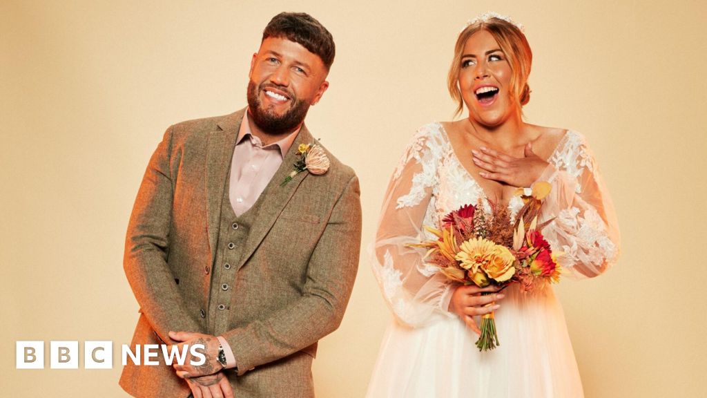 Polly and Adam in wedding outfits, with her wearing a white dress with sheer arms and a low v-neck and holding a bouquet of orange and red flowers while Adam is wearing a tweed suit in a pale brown with a pastel pink shirt and buttonhole.