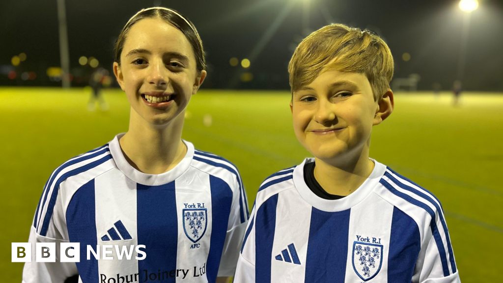 Two young girls wearing blue and white vertically striped football shirts are smiling at the camera.
There is a green football pitch behind them with floodlights.
