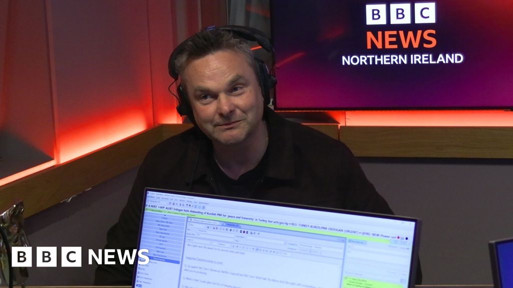 A man with headphones on in a radio studio. In the foreground, in front of him, we can see the front of a computer screen used by the radio programme presenter. In the background is a screen showing the BBC News Northern Ireland logo