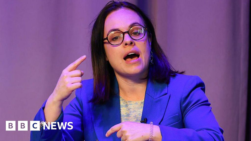 Kate Forbes, wearing small black round glasses and a blue blazer, she is mid speech and is gesturing with her index finger.
