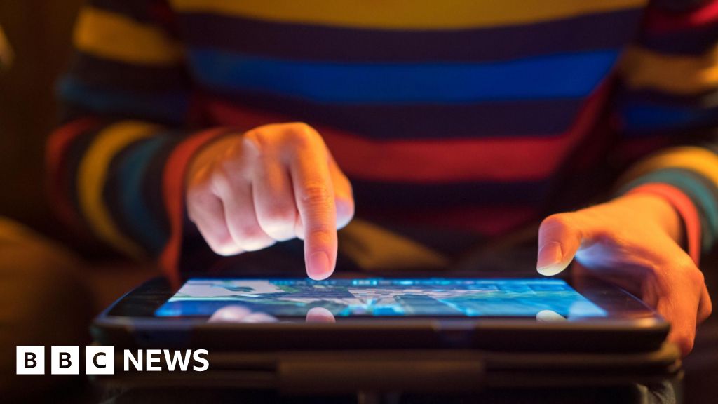 An image of a person using an iPad. It shows a child holding the device with two hands, one holding the iPad and another with one finger on the screen. The child is wearing a colourful stripy jumper