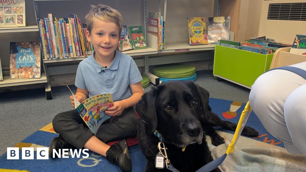 Seven-year-old Alfie sits on the floor next to Rafa, the black Labrador. He is reading him a book called Party Games. A book shelf with other children's books on it is behind Alfie and Rafa.