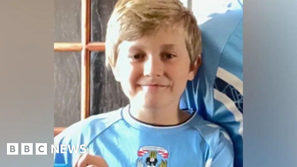 A boy in a sky blue Coventry City home shirt is smiling at the camera