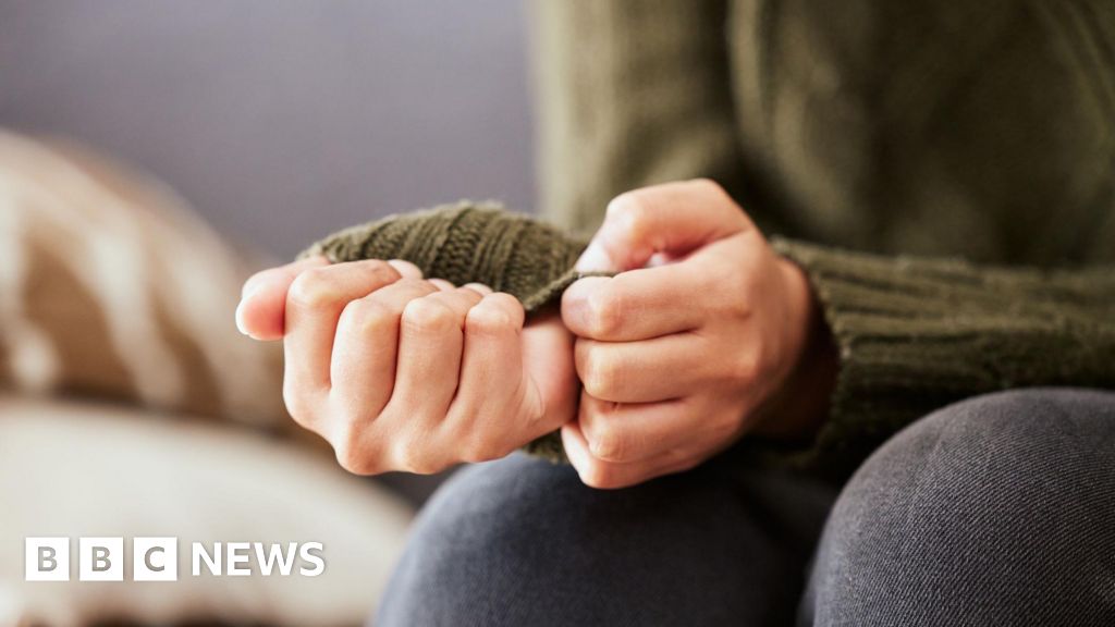 A woman sits on a sofa. She is pulling on a sleeve of her green jumper in a sign of feeling anxious.