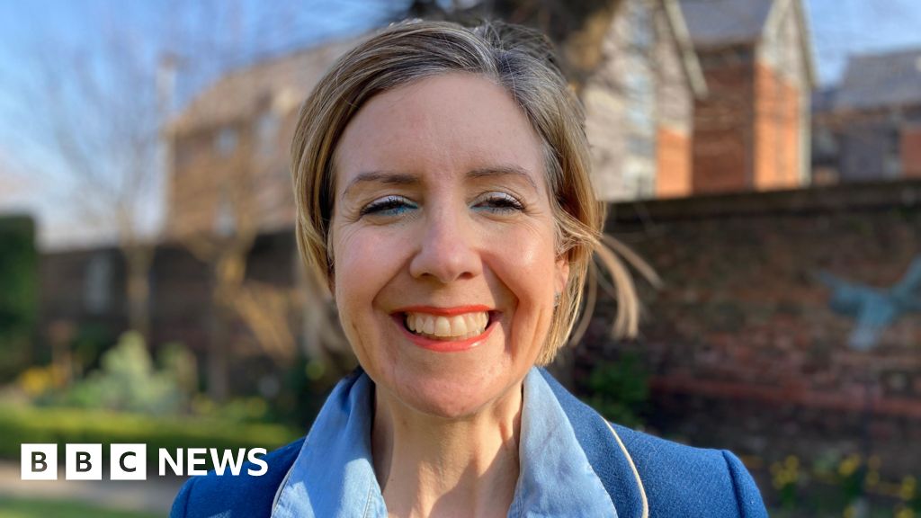 A head-and-shoulders photograph of Dame Andrea Jenkyns. She is standing in a garden and wearing a blue shirt and a blue jacket. She is smiling at the camera.