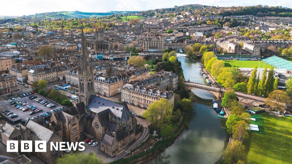 An aerial view of Bath city centre taken by a drone. Landmarks such as the River Avon and Bath Rugby's Rec stadium are visible and it is a clear, sunny day with a range of hills in the background