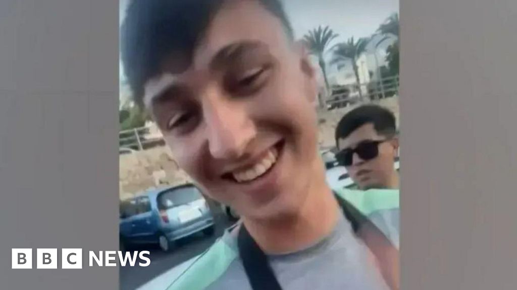 A picture of a smiling Jay Slater in a green and grey t-shirt with palm trees and a car in the background.