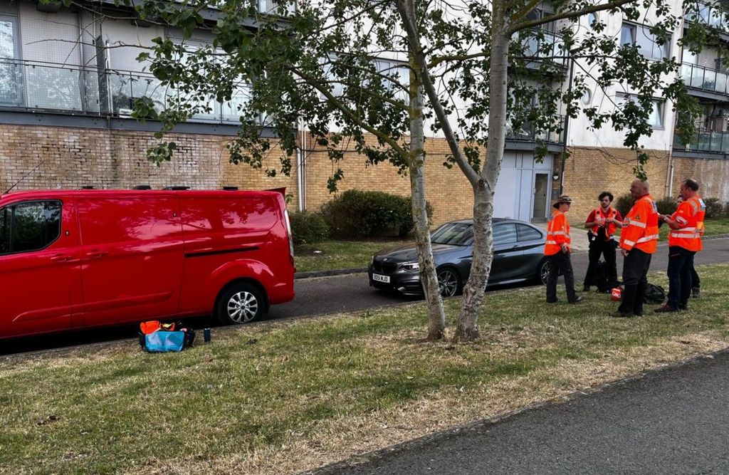 People, wearing orange jackets, stand on a grass verge in front of a block of flats  in Greenwich 