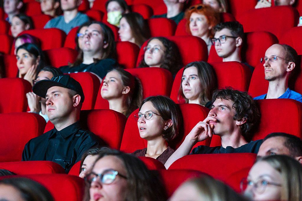 People sit in a cinema auditorium on rows of red chairs.