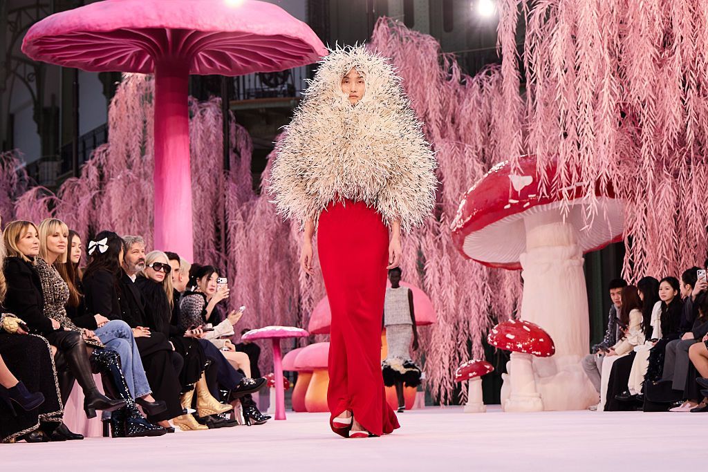 A model wearing a floor-length red dress with a cream fluffy jacket which cocoons her head, with her face poking out in the middle - there are massive pink toadstools and pink weeping willow trees behind her, and the audience sits watching on either side of the runway