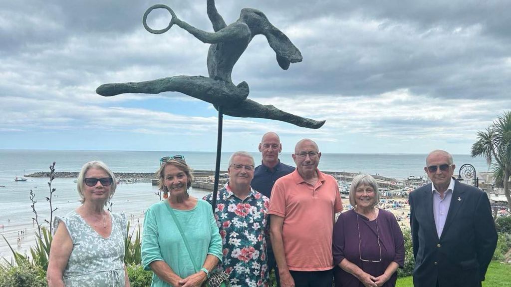 Three women and four men posing in front of a statue of a juggling hare. In the background is Lyme Regis seafront a the harbour