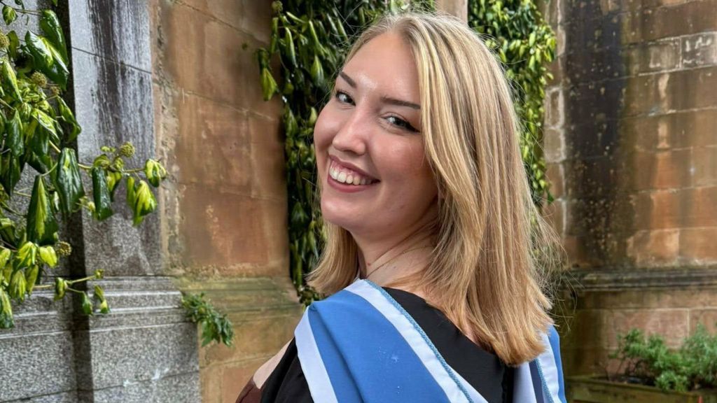 A young woman looking back over her shoulder wearing her black and blue graduation gown, against a backdrop of an old sandstone building and greenery.