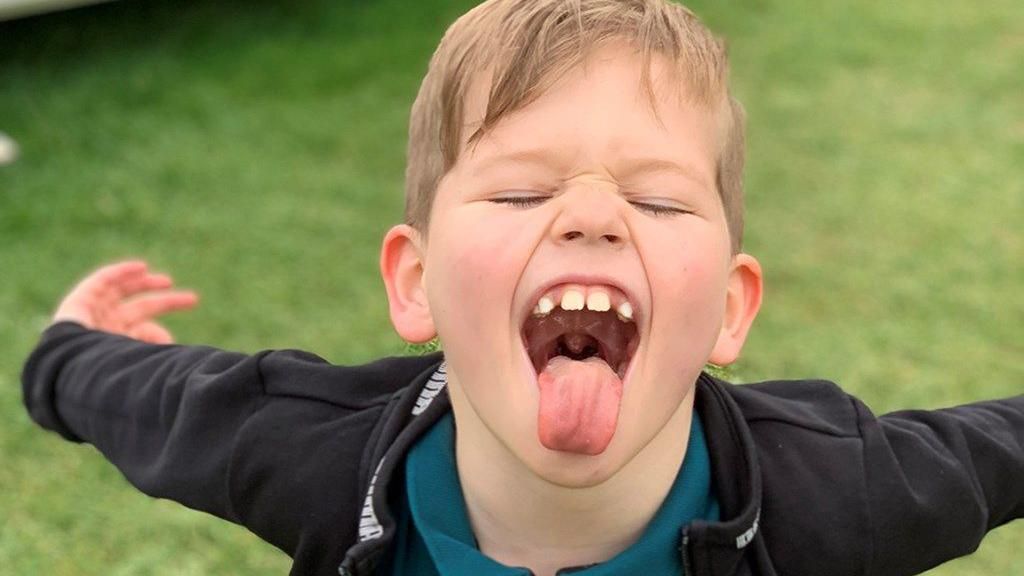 An eight-year-old boy with his arms outstretched behind him, his eyes squeezed shut and his tongue sticking out in a playful manner