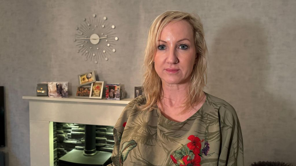 A picture of Aimee Geary, with long blonde hair and wearing a green top, standing in front of a fire place