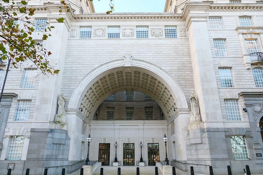 The grand stone entrance of Thames House, the London headquarters of MI5, showing a large arched gateway, carved stonework and glass-panelled doors, with black security bollards lining the pavement in front.