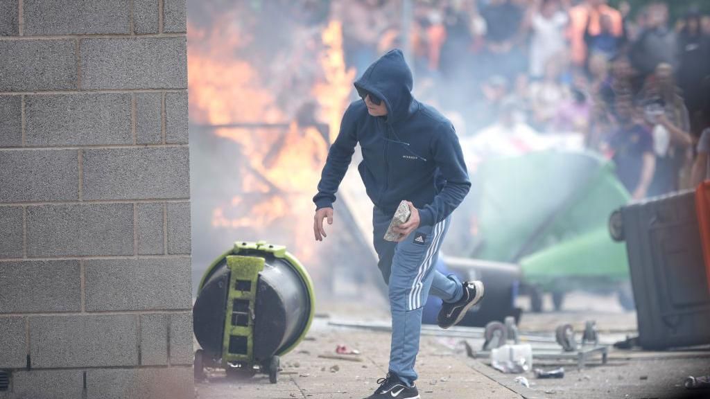 A man holds a brick with a fire and scenes of disorder in the background.