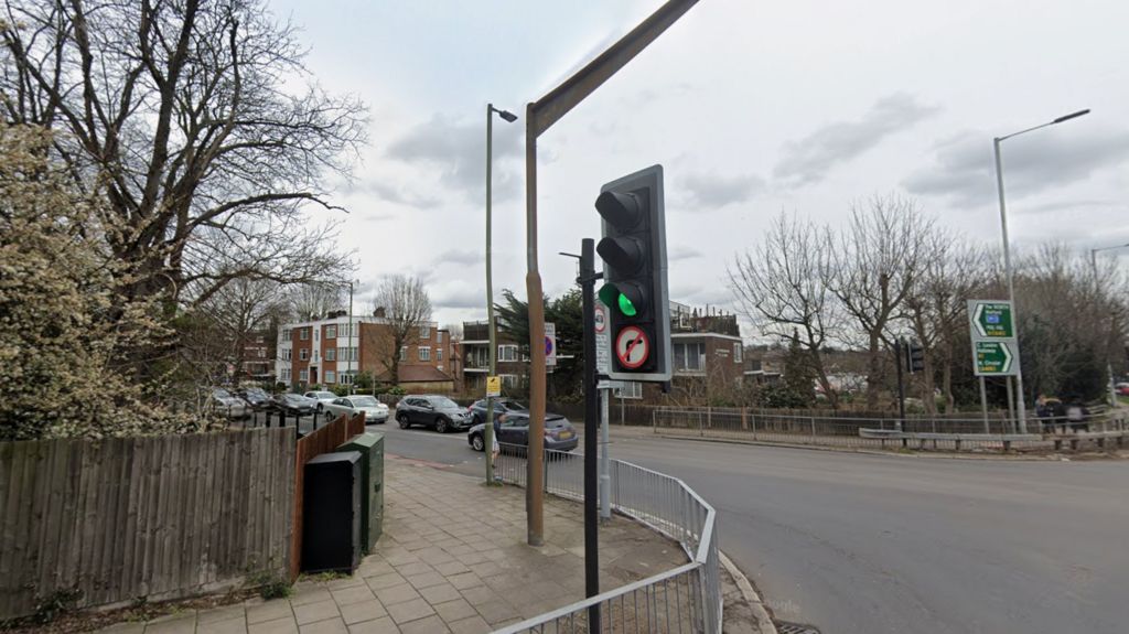A general view of a pedestrian traffic light crossing on Holders Hill Road, with cars on the road and residential buildings and trees in the background.