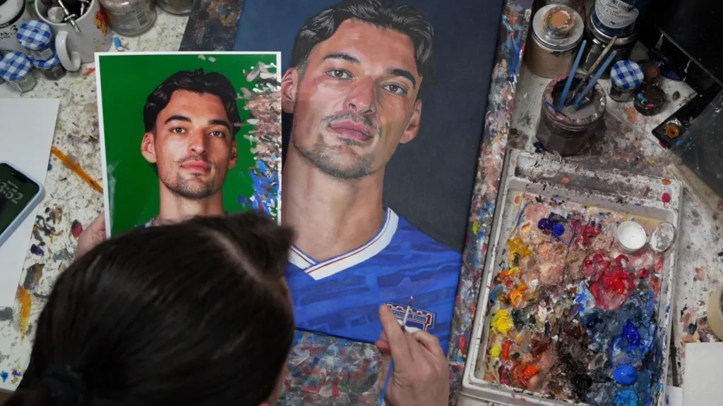 An overhead shot of Louise Cobbold, showing the top of her head, with its dark brown hair pulled back. She is looking at a paint splattered table where thre is a photo of Jacob Greaves alongside her painting of him. To the right is a tray full of smudged acrylic paint. 