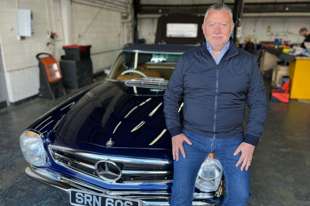 Justin Linny perched on the bonnet of a classic blue Mercedes Benz in a car workshop 