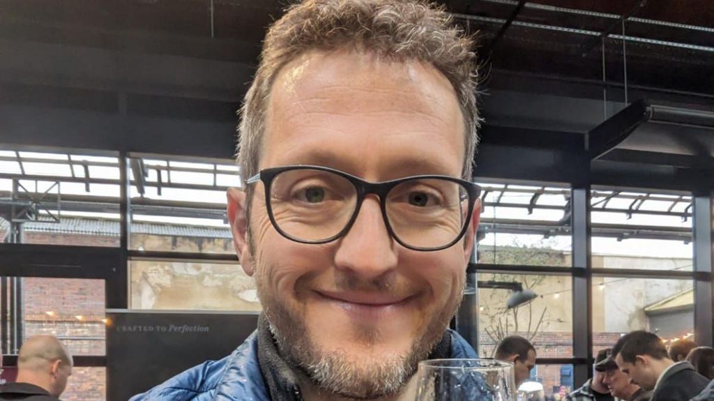 Peter Walker wearing spectacles while waiting in Tenereife airport. He is smiling and has a short beard and short, light brown hair.