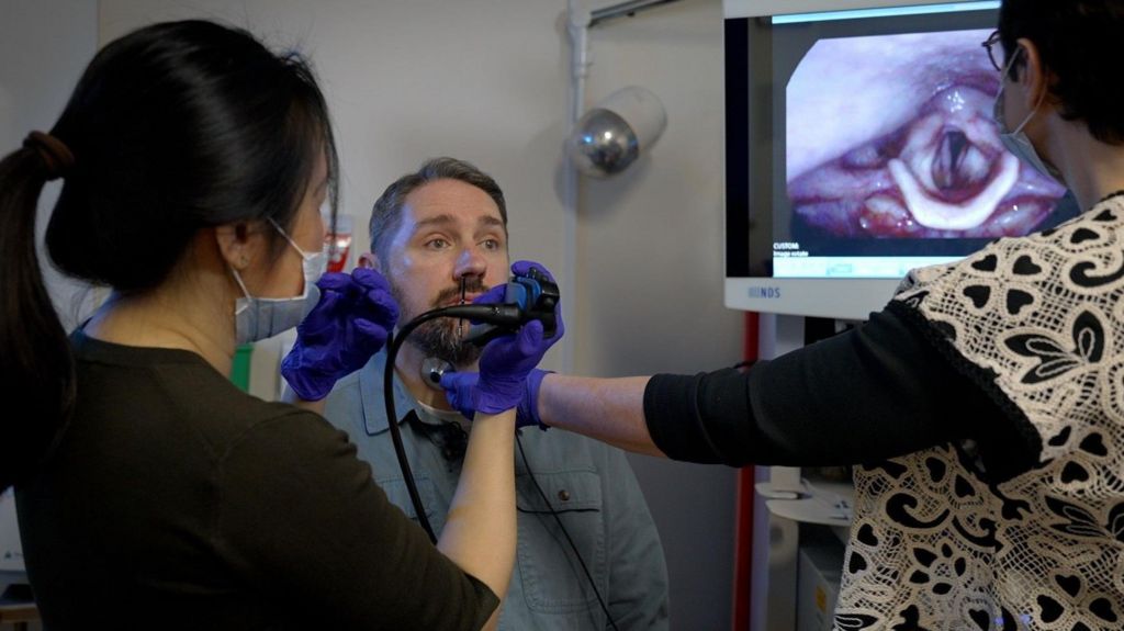 A man sits in a chair as his doctor and speech therapist view images of his vocal cords on a screen