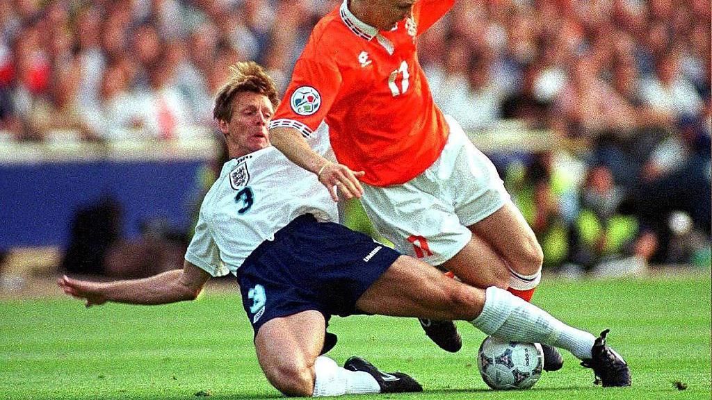 Stuart Pearce, who is wearing a white England kit, has his leg outstretched and is tackling a player in a red kit. They are on a football field.